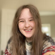 Headshot of Milo Machin smiling at the camera. They are pale skinned with long brown hair and are wearing a knitted jumper/sweater